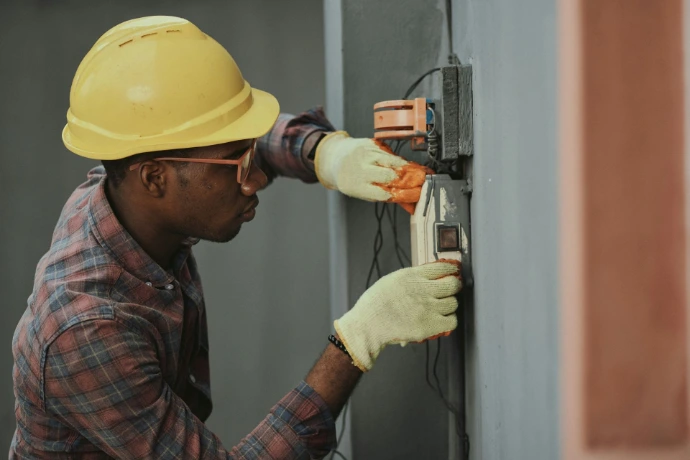 Person wearing a hard hat and safety gloves inspecting an electrical switch on a wall